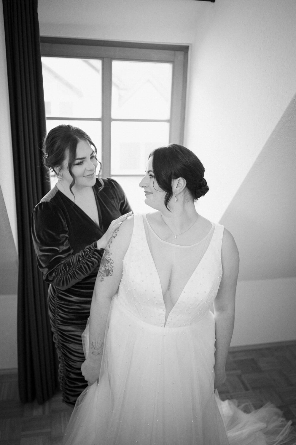 Bride smiling while friend helps with wedding dress during morning preparations.