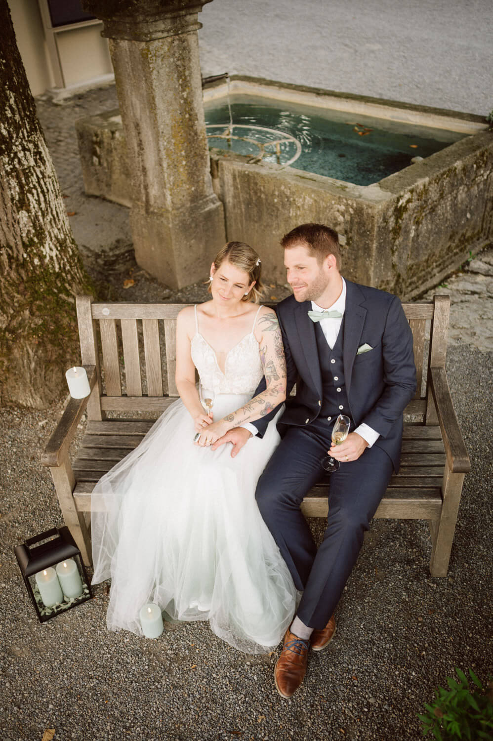 Bride and groom sitting on a wooden bench with champagne near a stone fountain, luxury wedding photography.