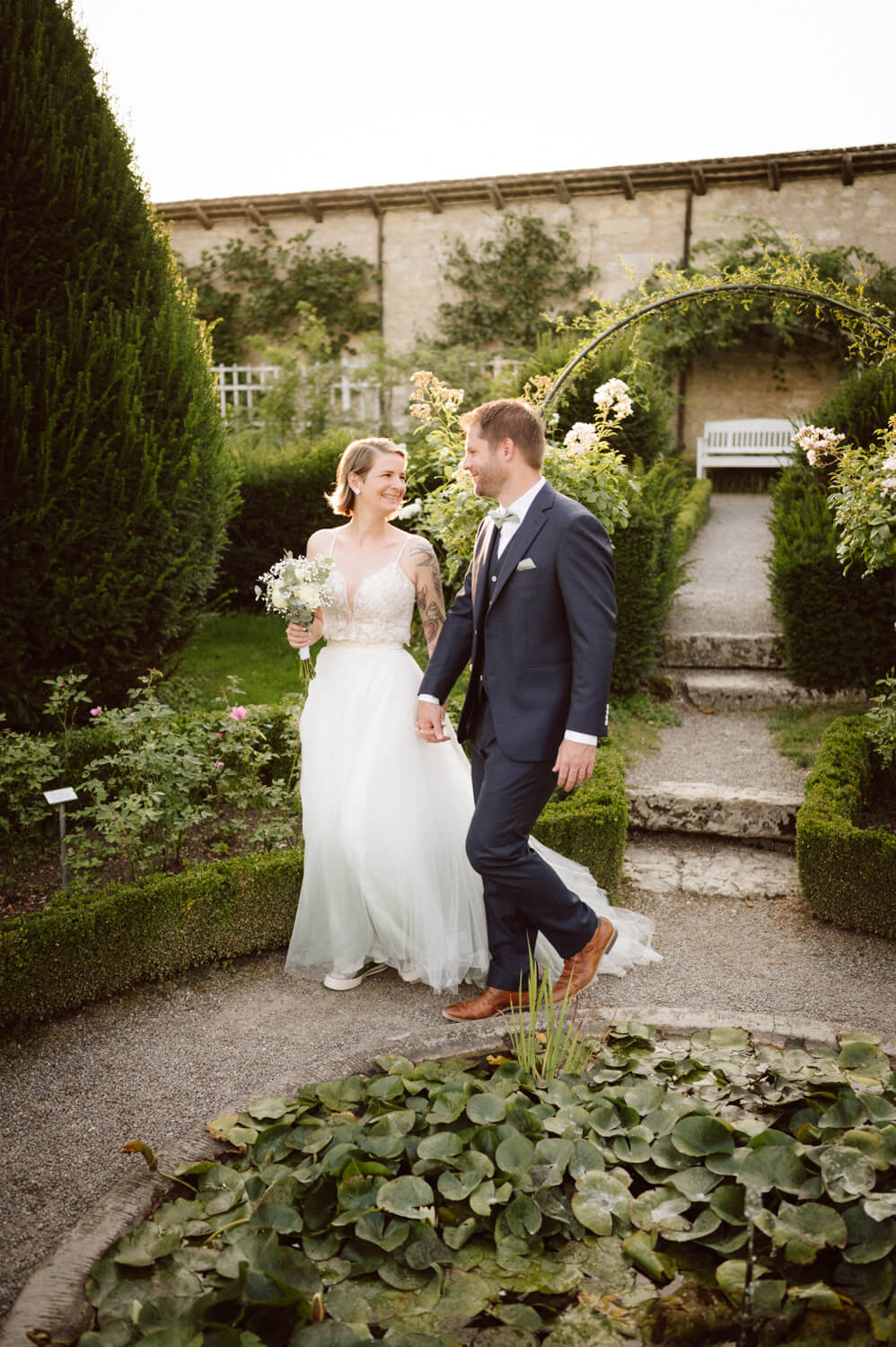 Bride and groom walking hand in hand through a rose garden, smiling and joyful, luxury wedding photography.