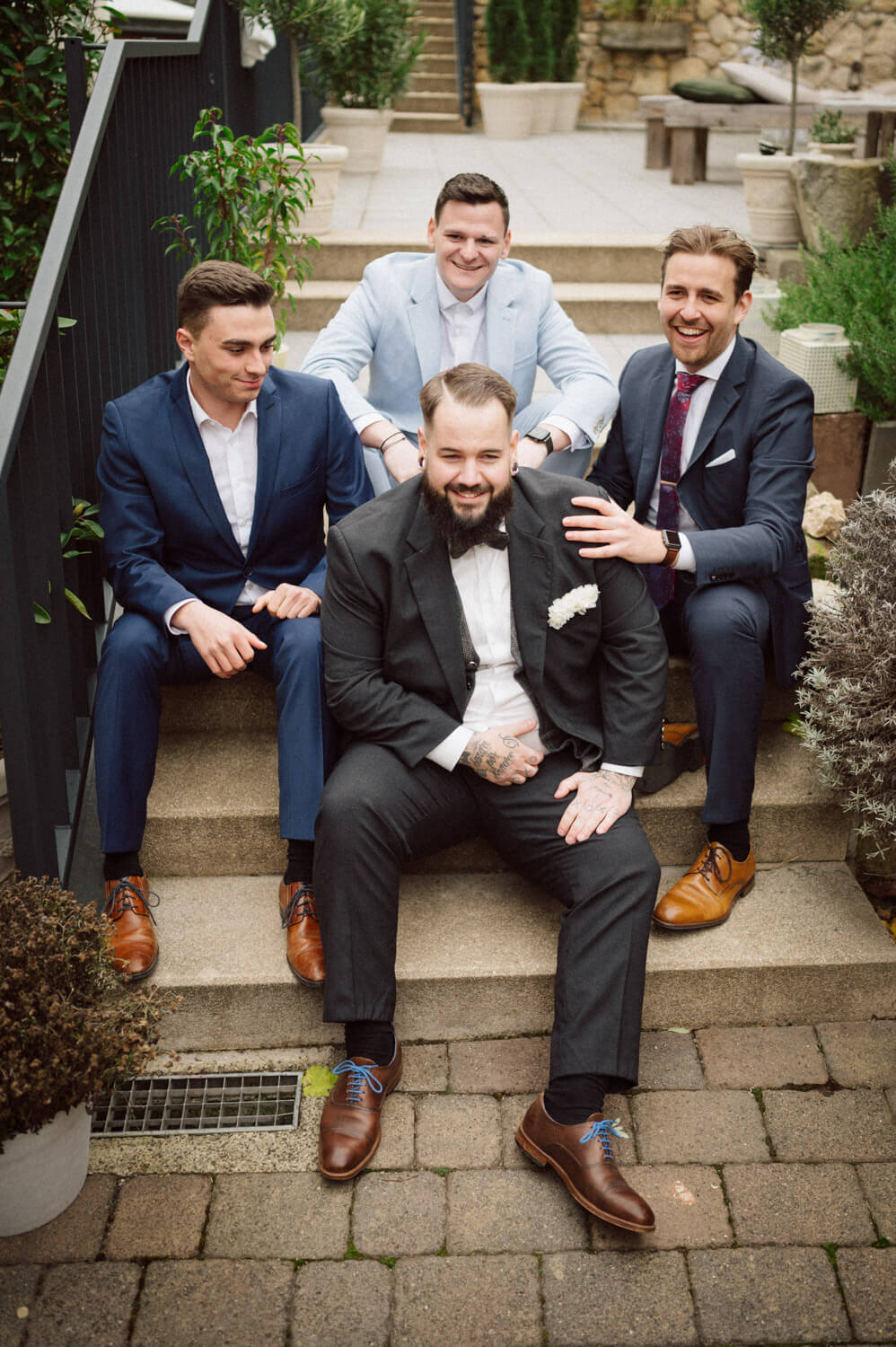 Groom sitting with his groomsmen on outdoor stairs, smiling in elegant suits, luxury wedding photography.