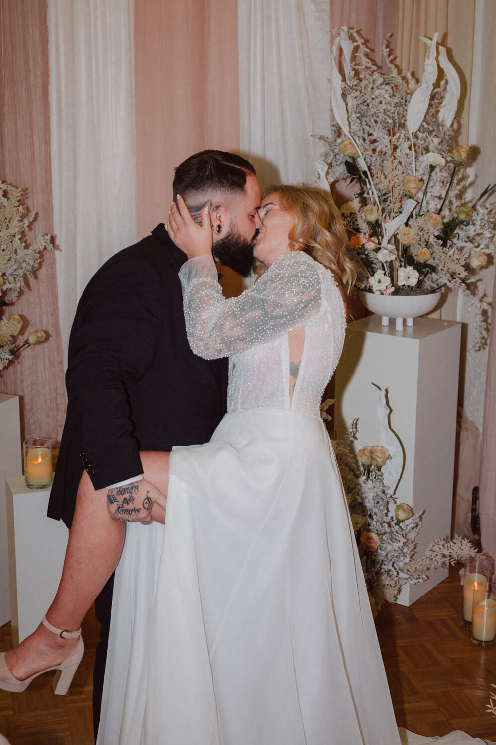 Bride and groom sharing a passionate kiss indoors with floral decoration and candles, luxury wedding photography.