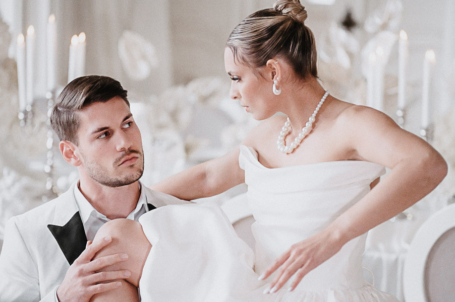 ditorial wedding portrait of a couple in a white ballroom—bride in an off-shoulder gown with pearl necklace and earrings, groom in a white tuxedo, candles in the background.