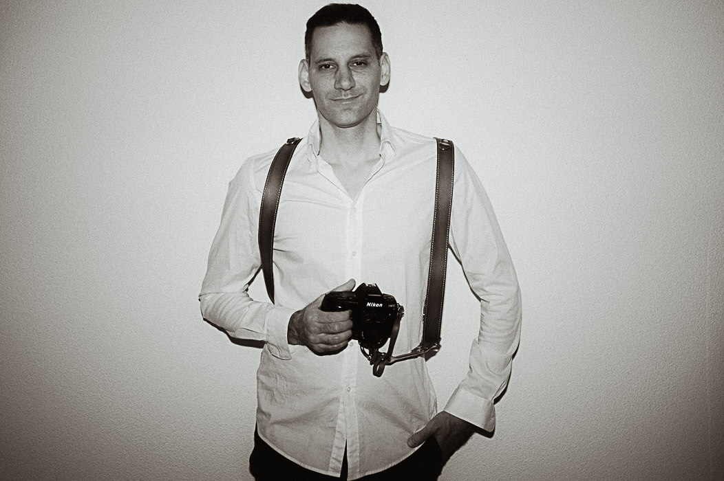 Black-and-white portrait of a wedding photographer holding a Nikon camera with a leather shoulder harness, standing against a plain wall.