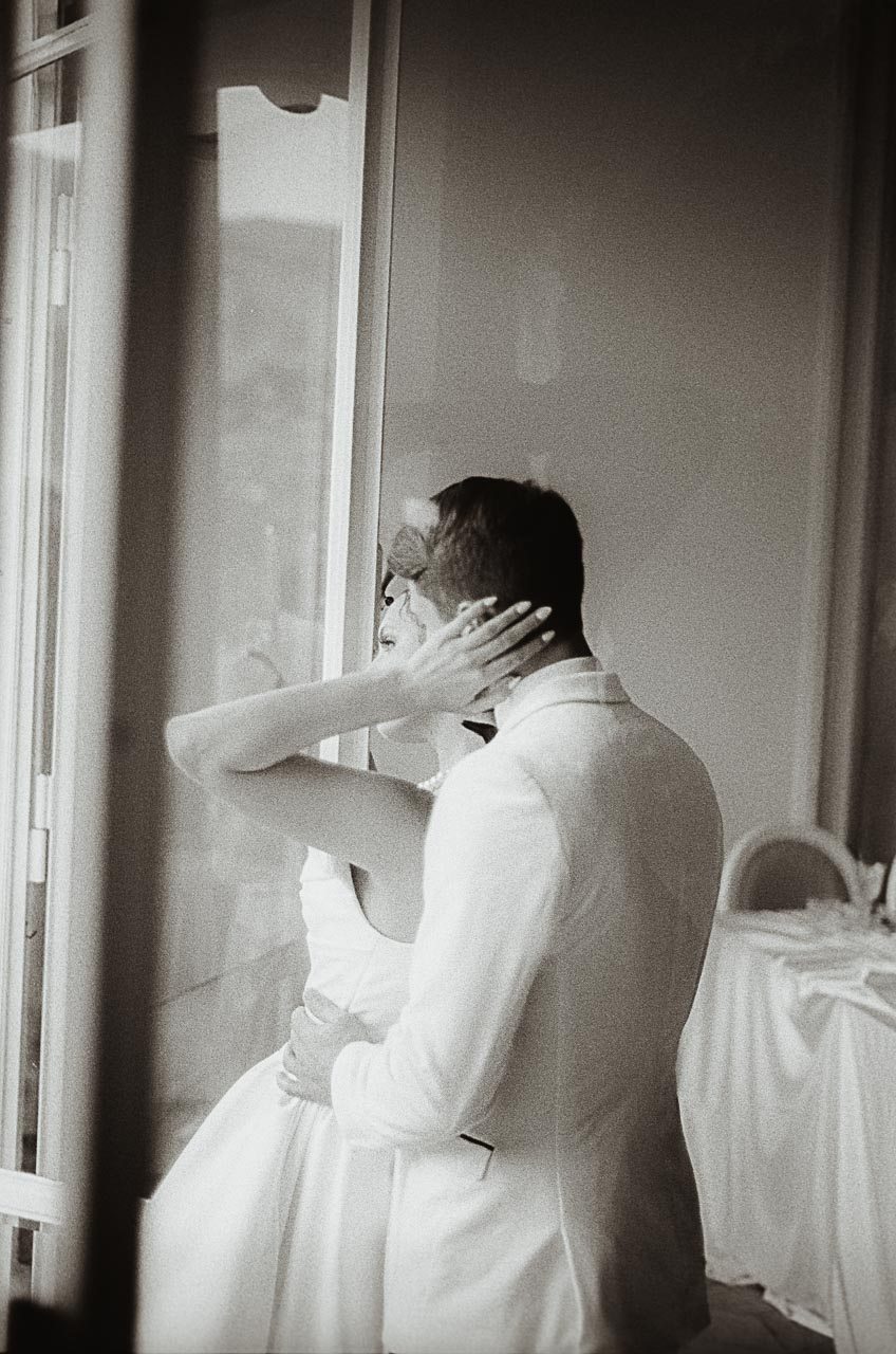 Backlit embrace by the window, bride touching the groom’s face, photographed on Kodak Tri-X film.